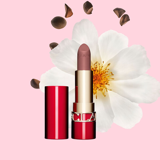 Packshot of Clarins matte Lipstick Joli Rouge in Shade 795 V, placed beside a camellia flower and shea seeds on a pink background.