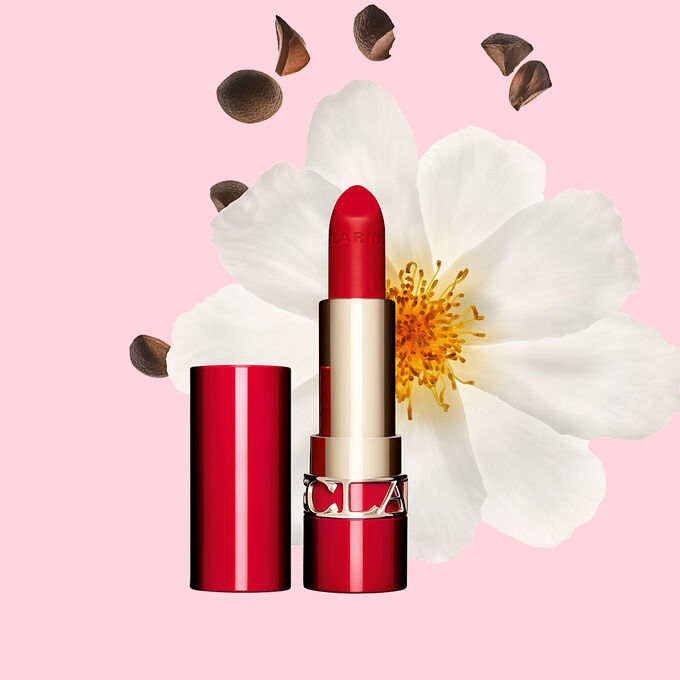 Packshot of Clarins matte Lipstick Joli Rouge in Shade 768 V, placed beside a camellia flower and shea seeds on a pink background.