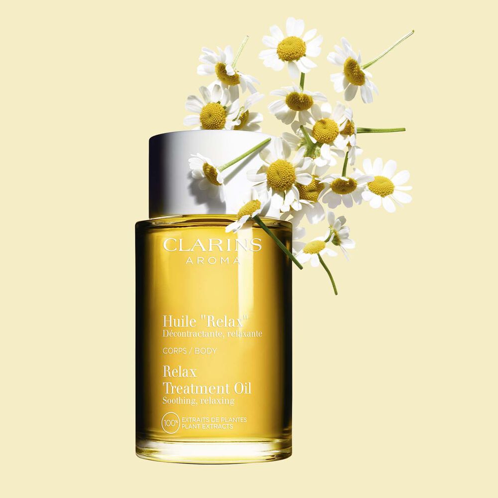 Packshot of a bottle of Clarins Relax Body Treatment Oil close to a bouquet of chamomile flowers on a pale yellow background.