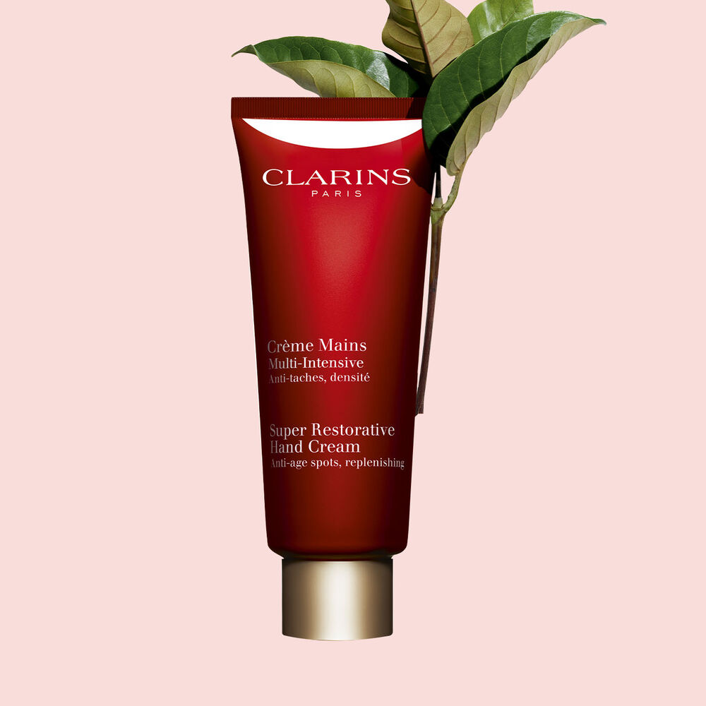 Packshot of a bottle of Clarins Super Restorative Hand Cream close to some harungana leafs on a pink background.