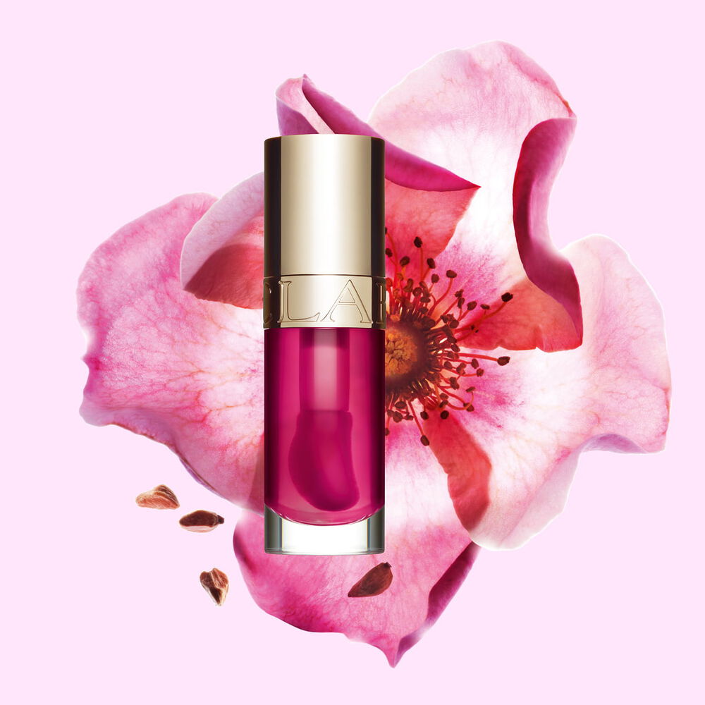 Packshot of Clarins Lip Comfort Oil in Raspberry, placed next to a sweetbriar rose on a light peach background.