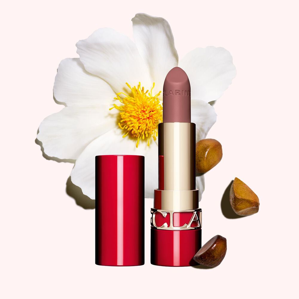 Packshot of Clarins matte Lipstick Joli Rouge in Shade 795 V, placed beside a camellia flower and shea seeds on a pink background.