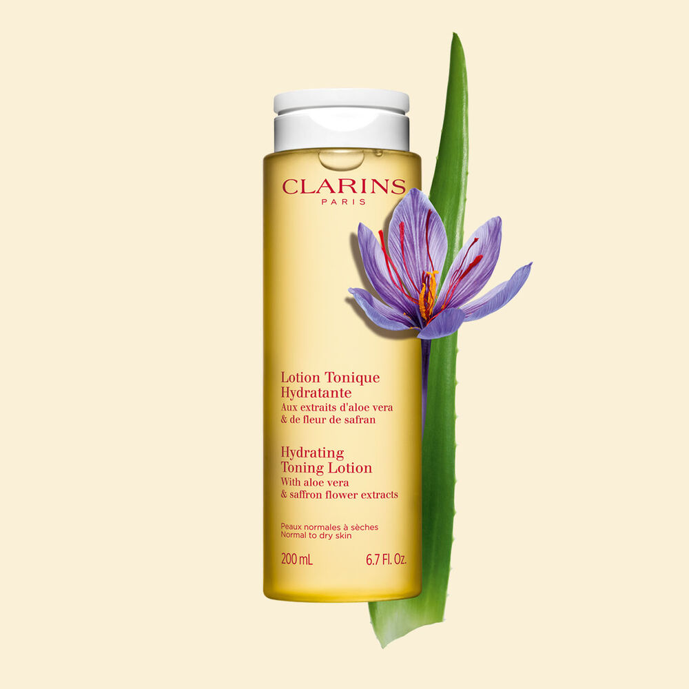 Packshot of the face lotion yellow bottle next to a saffron flower and a piece of aloe vera on a yellow background