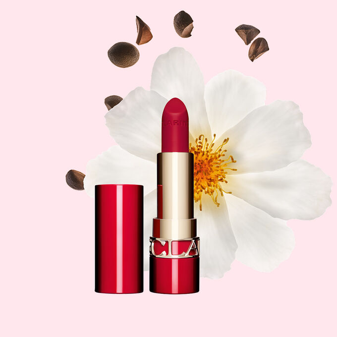 Packshot of Clarins matte Lipstick Joli Rouge in Shade 742 V, placed beside a camellia flower and shea seeds on a pink background.