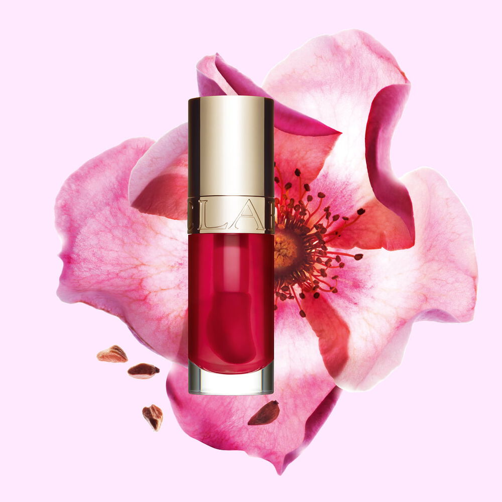 Packshot of Clarins Lip Comfort Oil Fushia, placed next to a sweetbriar rose on a light pink background.