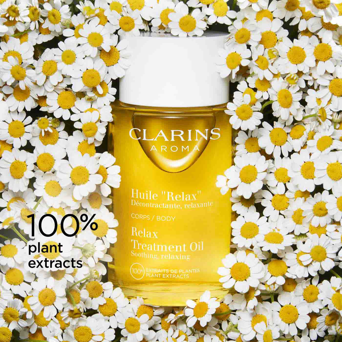 Bottle of Clarins Relax Body Treatment Oil among chamomile flowers to illustrate it's made of 100% plant extracts.